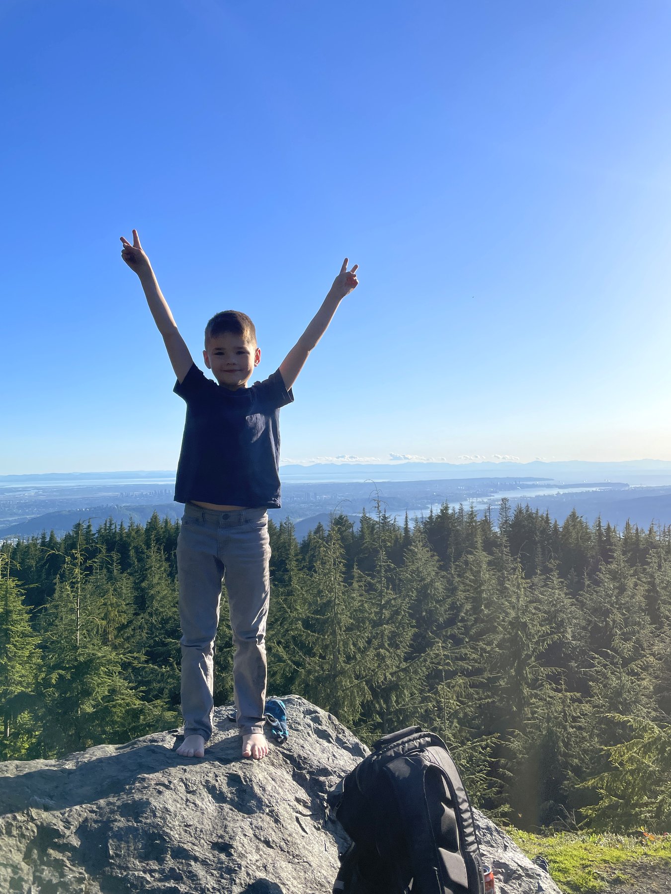 Craig Johnston's son at the summit of Eagle Mountain in Coquitlam, BC, arms raised in a peace-sign celebration with a view of the Lower Mainland and water in the distance behind him.