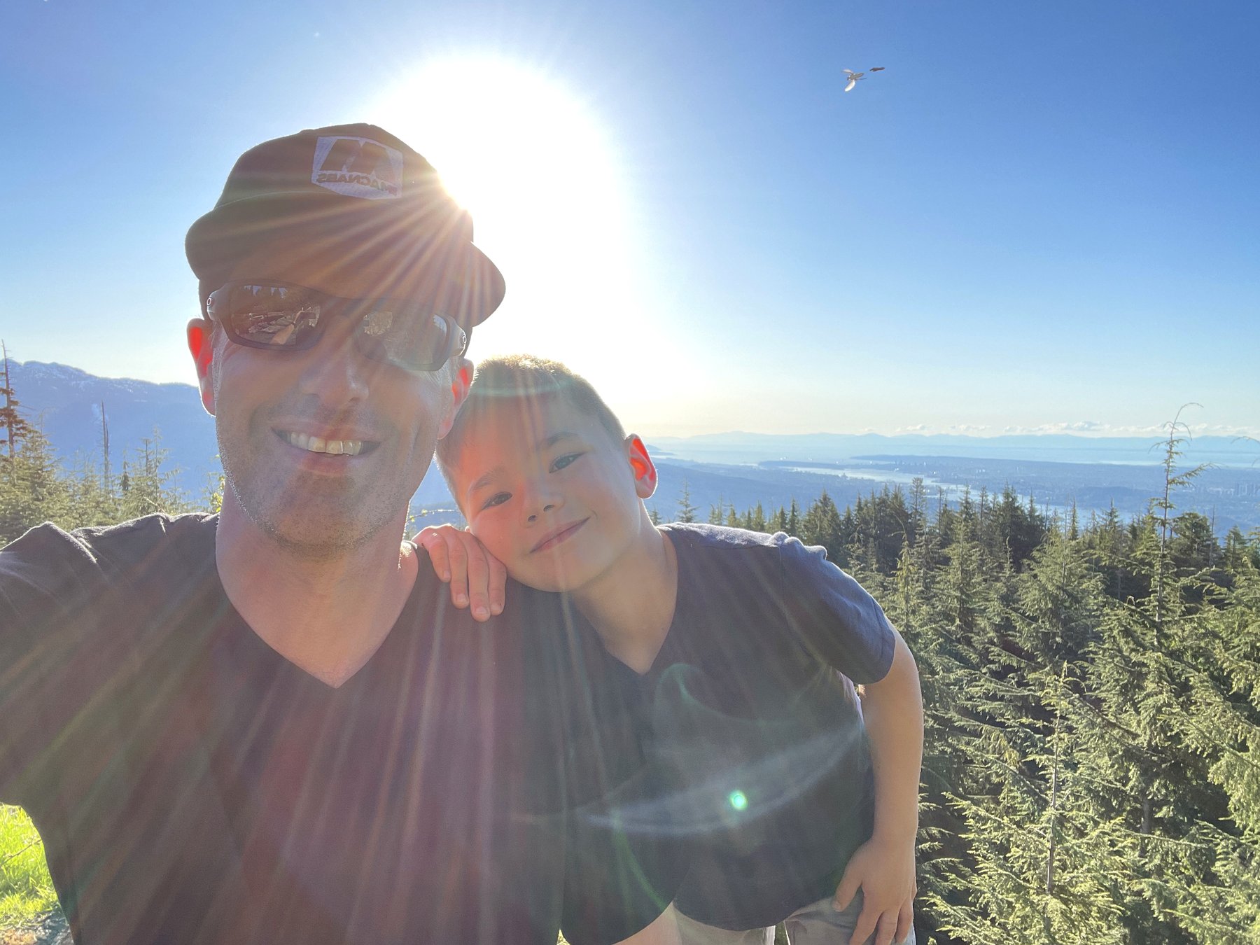 Craig Johnston and his son at the summit of Eagle Mountain in Coquitlam, BC, with the Burrard Inlet and Lower Mainland visible behind them. Craig is wearing a MACNABS hat. A father-son afternoon on the mountain that buyers cannot see from a listing photo.